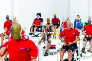 Rows of crash test dummies in red T-shirts, some seated on test rigs and vehicle seats, others fitted with exposed mechanical torsos and jointed limbs, lined up inside Geely's Safety Center testing facility